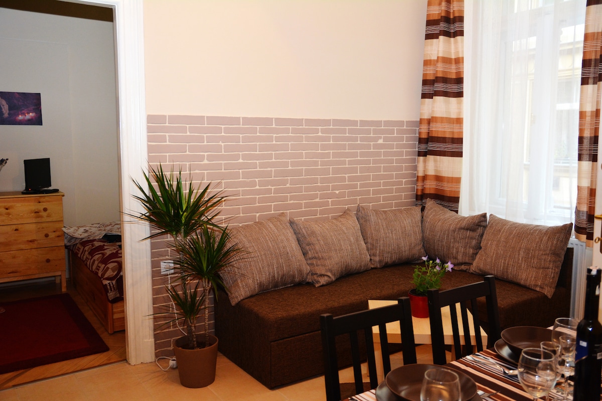 A spacious seating area is highlighted by a comfortable sectional sofa adorned with neutral-hued cushions. A potted plant adds a touch of greenery beside the sofa. A dining table is set with glasses and cutlery, creating an inviting space for meals.