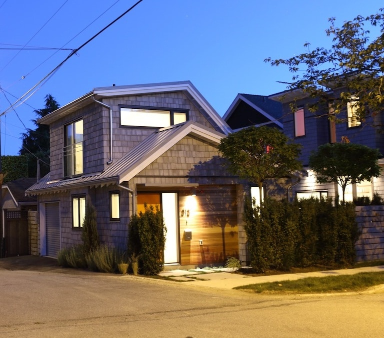 A modern coach house exterior is shown at dusk, featuring a wooden door and large windows illuminated softly. The building is framed by landscaped hedges and is situated at the corner of a quiet street, with neighboring properties partially visible.