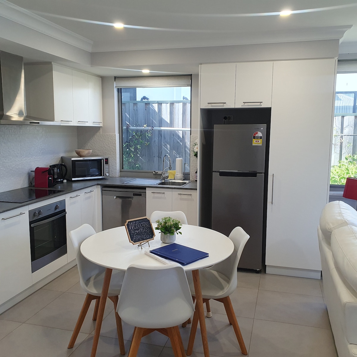 A modern kitchen area is presented, featuring sleek cabinetry and a polished countertop. A round dining table with white chairs occupies the space, accompanied by stainless steel appliances and natural light from the nearby window, enhancing the inviting kitchen atmosphere.