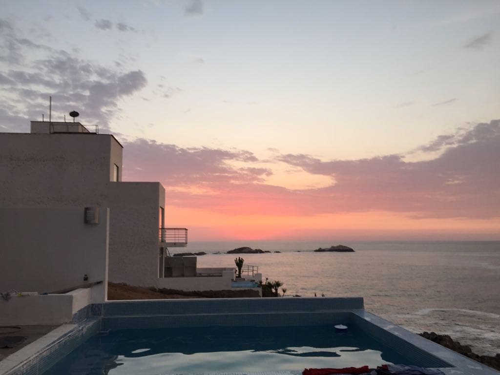 The image captures a tranquil evening sky, transitioning from soft pink to deep blue as the sun sets over the ocean. A swimming pool is visible in the foreground, reflecting the vibrant colors of the sunset, while the modern structure of the house stands nearby.
