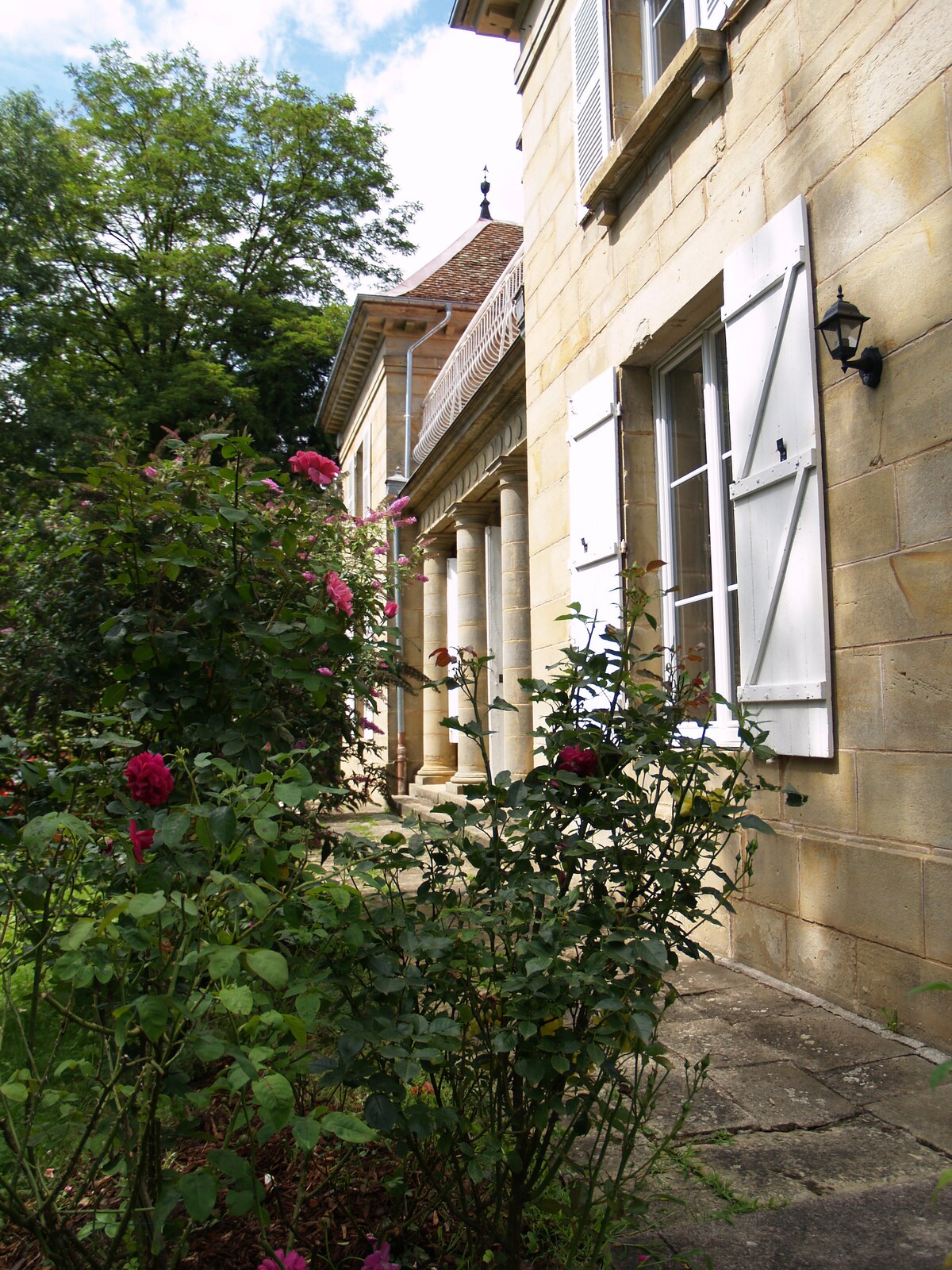 The image captures a charming exterior view of the château, featuring light-colored stone walls and white shutters. Lush green shrubs and blooming pink roses are positioned in the foreground, enhancing the natural beauty of the surroundings while tall trees provide a serene backdrop.