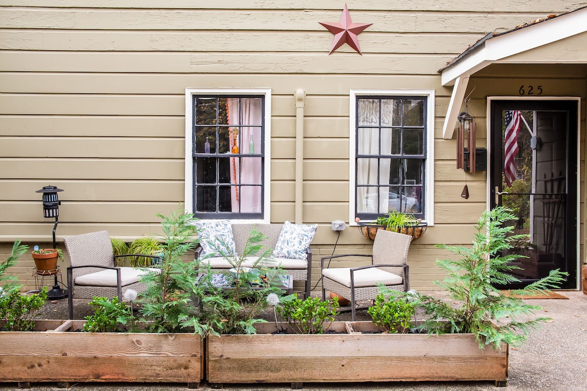 An inviting front patio showcases a two-seat bench and two individual chairs, framed by planters with greenery. A star decorates the wall above, and window treatments allow soft light to enter the space. A walkway leads to the entrance of the upstairs apartment.