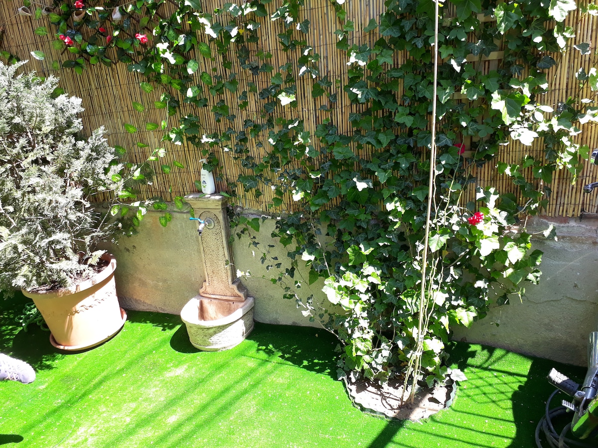 A private courtyard is filled with vibrant green artificial grass, surrounded by climbing ivy and potted plants. A decorative fountain is positioned against a textured wall, enhancing the tranquil outdoor space. Sunlight filters through, creating a peaceful atmosphere.