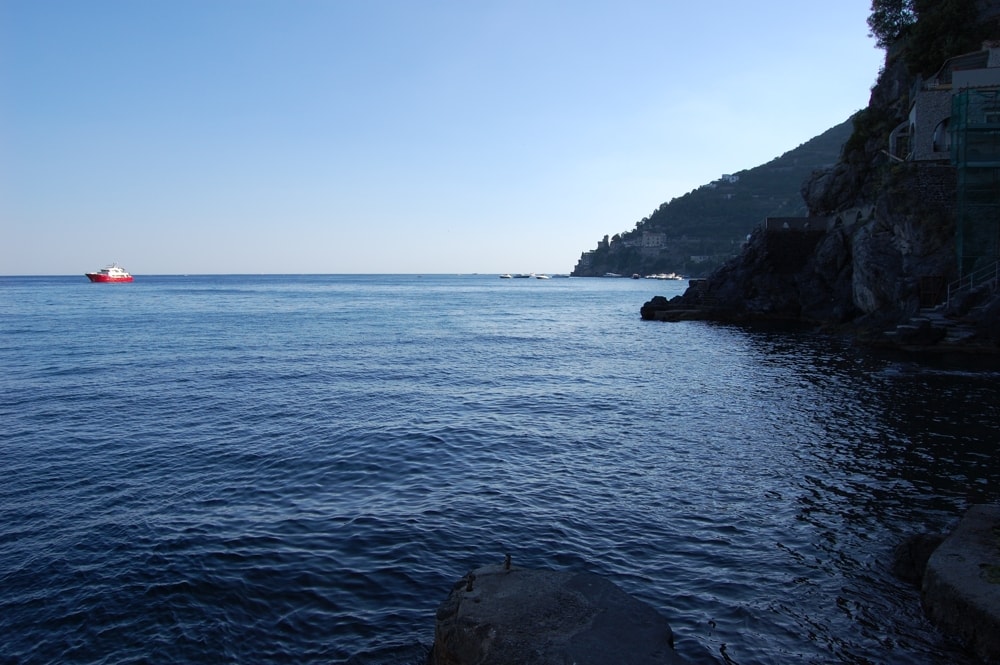 A serene view of the calm sea, with gentle waves reflecting the clear blue sky. A small boat is seen on the water, while the rocky shoreline extends towards a hillside dotted with greenery and distant buildings.