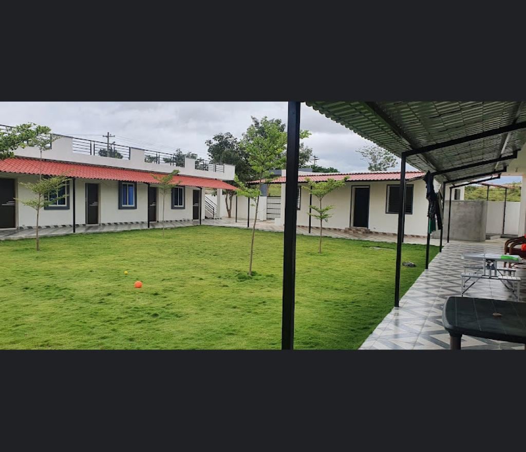 A grassy courtyard is bordered by single-story buildings with red roofs and white walls. The area features young trees and colorful ground markers scattered on the lawn. A covered patio with a tiled floor offers places to sit and relax, promoting outdoor enjoyment.