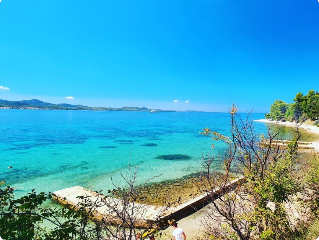 A serene coastal view presents clear blue waters meeting the horizon, with gentle waves lapping against a rocky shoreline. Green vegetation frames the scene, while several people can be seen enjoying the refreshing water in the distance.