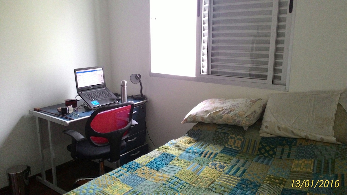 A spacious room features a cozy bed with a patterned bedspread and two pillows. A desk with a laptop and a chair is positioned beside the window, allowing natural light to filter through. A small wastebasket is placed next to the desk.