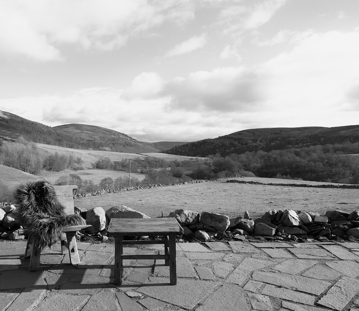 A rustic outdoor seating area with a wooden chair adorned with a faux fur blanket, accompanied by a small table. The expansive view showcases rolling hills and a clear sky, framed by a stone wall, providing a sense of peace and connection to nature.