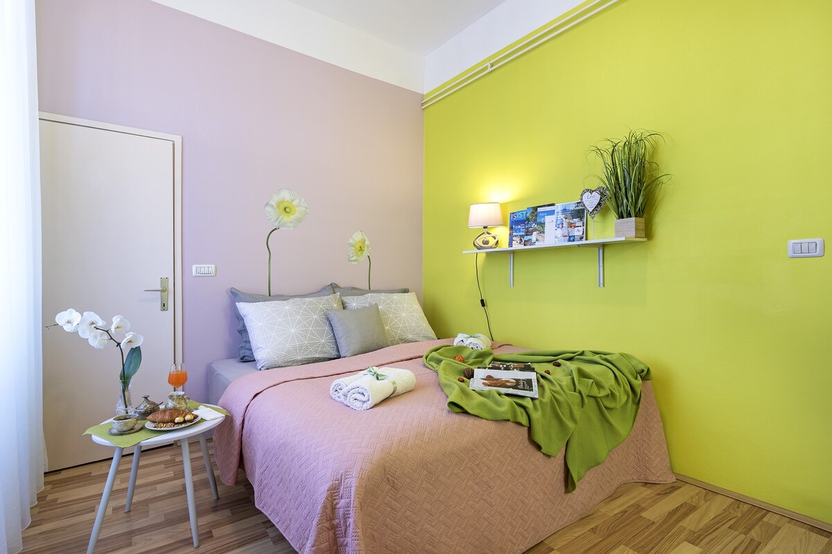 A cozy sleeping area is presented, featuring a double bed with layered blankets in soft pink and green tones. Bright green walls are complemented by pale pink accents and decorative wall shelves. A small table holds a beverage and snacks, inviting relaxation.