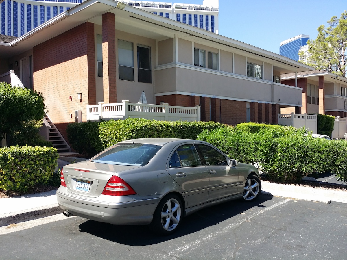 A silver sedan is parked in front of a two-story building surrounded by well-maintained greenery. The brick exterior of the building is complemented by white railings and staircases leading to upper units. Clear skies provide a bright backdrop, enhancing the clean and orderly environment.