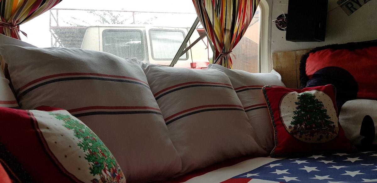 The image features a cozy seating area within the Airstream, adorned with plush pillows in red, white, and blue stripes, creating a comfortable ambiance. Festive throw pillows include circular designs with Christmas trees, contributing to the unique decor. Natural light filters through colorful striped curtains.