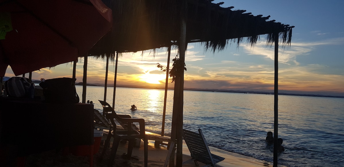 A serene beach scene captures a vibrant sunset over calm waters. A thatched roof provides shade over simple seating arrangements, with silhouettes of individuals in the water. The sky displays a blend of warm colors, creating a tranquil atmosphere reflective of nature's beauty.