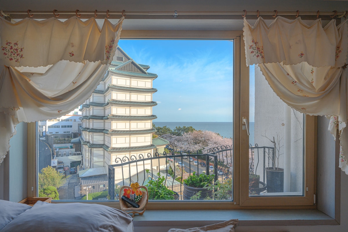 A window view showcases a serene ocean scene, framed by soft, decorative curtains. A vibrant cherry blossom tree can be seen nearby, with a neighboring building's unique architecture prominently displayed.
