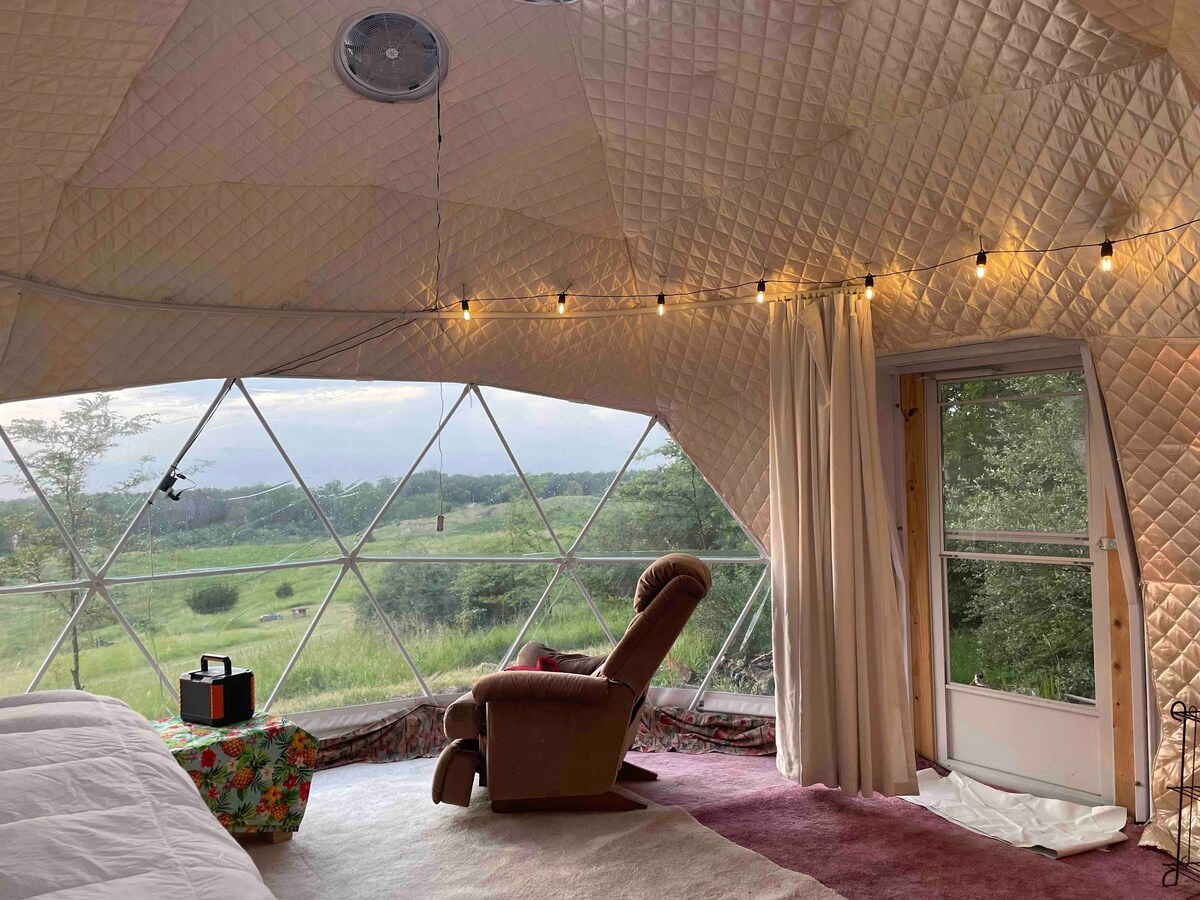 The interior of the geodesic dome features a cozy seating area with a recliner chair, set against a backdrop of large triangular windows that frame the expansive views of the surrounding nature. String lights provide soft illumination along the ceiling.