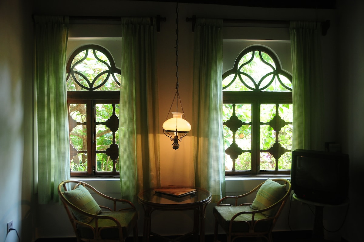 Two windows with decorative arches are framed by light green curtains, allowing natural light to illuminate the space. A cane table stands between two cozy chairs, with a pendant light hanging above. Lush greenery is visible outside, enhancing the serene ambiance.