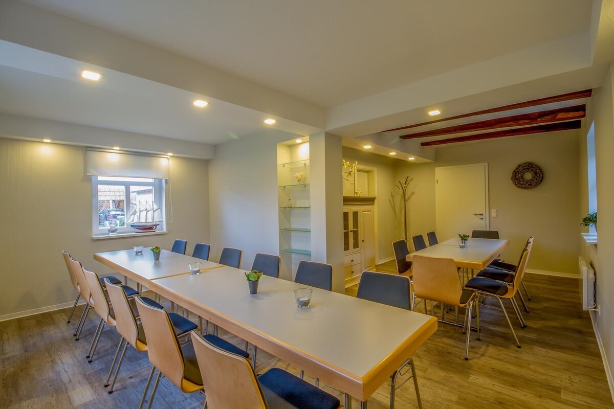 A spacious meeting room is shown, featuring a long table surrounded by multiple chairs. Soft lighting is provided by recessed ceiling fixtures, and a large window allows natural light to fill the space. Shelving units add a functional touch to the room.
