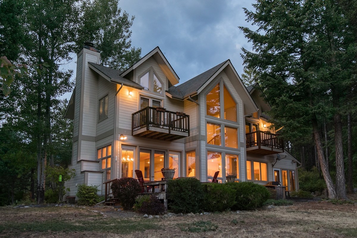 The exterior of a modern home is presented, showcasing large windows that provide beautiful views and warm lighting. A deck with seating invites relaxation. Surrounding trees and shrubs offer a natural setting, emphasizing the peaceful atmosphere of the property.