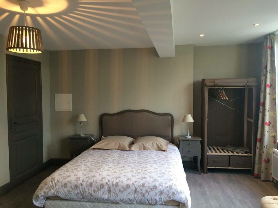 A spacious sleeping area features a large bed with light-toned bedding, complemented by two bedside lamps on dark nightstands. A decorative light fixture casts a patterned shadow on the ceiling. A wardrobe with hanging space is positioned nearby, and light enters through a window draped with floral curtains.