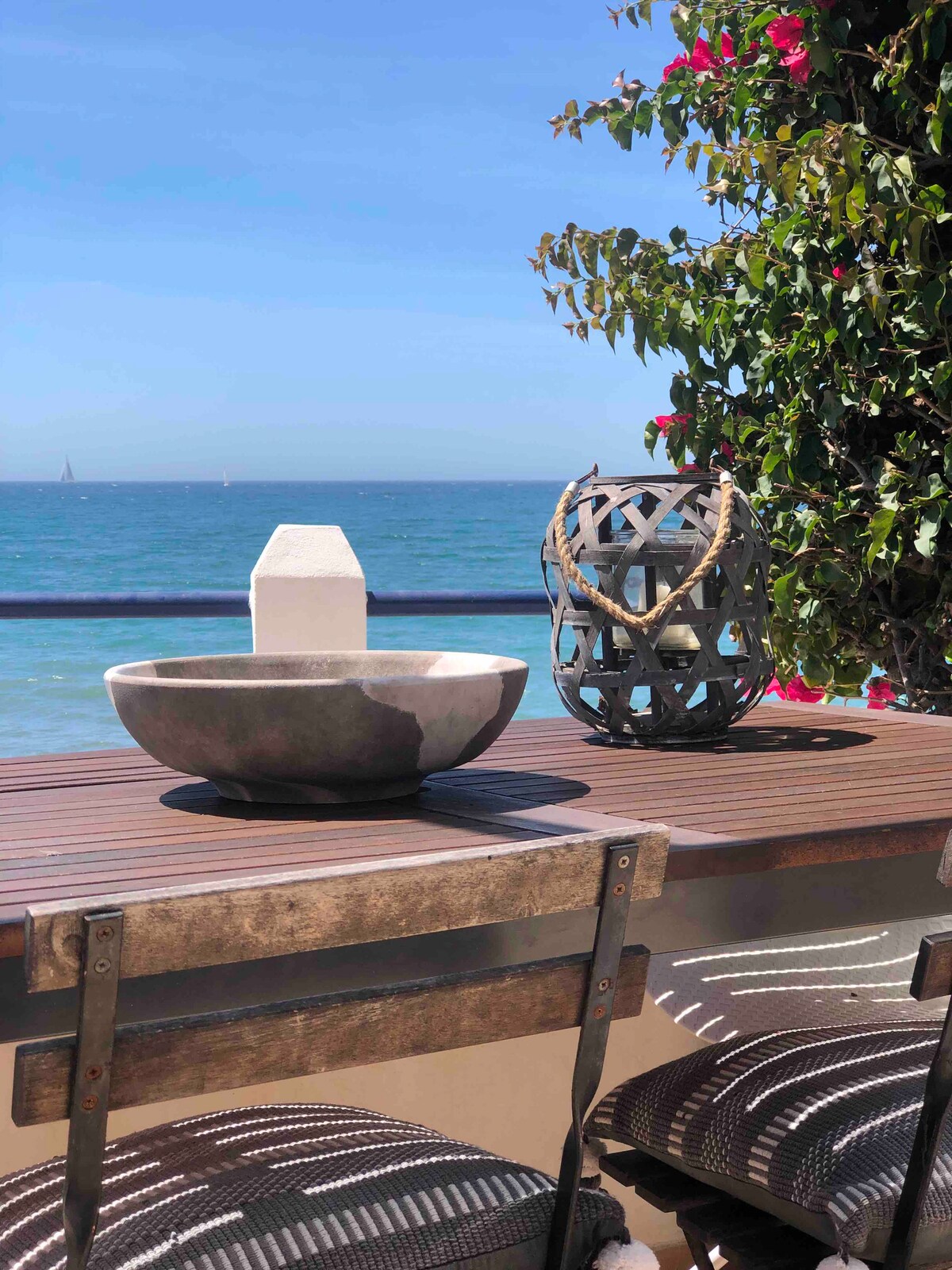 A wooden dining table is positioned by the sea, adorned with a round stone bowl and a textured lantern. Vibrant flowers climb along the railing, with the ocean and a clear sky visible in the background.