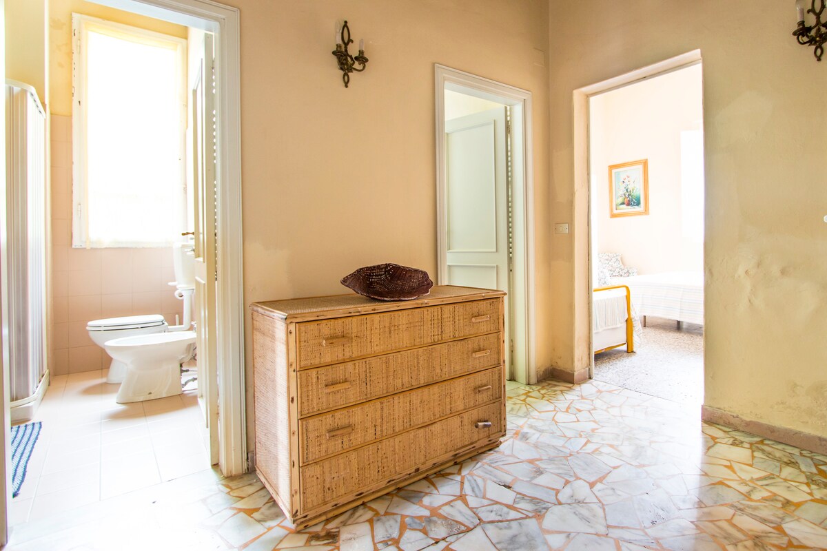 The image displays a spacious entryway featuring a wooden storage chest and light-colored walls. Doorways lead to a bathroom on the left and a bedroom on the right, with natural light filtering in from a nearby window. A tiled floor adds a subtle texture to the setting.