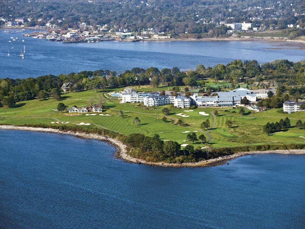 An aerial view captures the lush landscape of the resort, featuring expansive green golf courses and buildings nestled along the coastline. The beautiful Penobscot Bay is visible in the background, showcasing calm waters and a distant shoreline dotted with boats and homes.