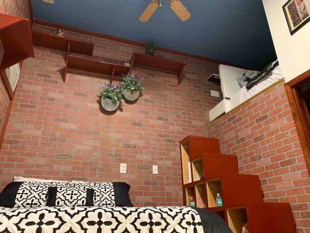 A cozy room features exposed brick walls, complemented by wooden shelves adorned with potted plants. A spacious bed is dressed in a patterned black and white blanket, while a staircase leads to an elevated sleeping area. A ceiling fan is visible above.