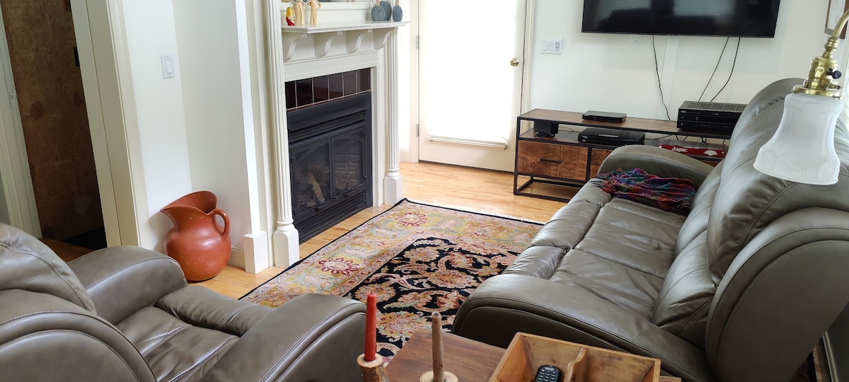 A living area features leather seating arranged around a decorative area rug. A gas fireplace is centered beneath a mantel, with a flat-screen TV mounted on the wall. Natural light enters through glass doors leading outside.