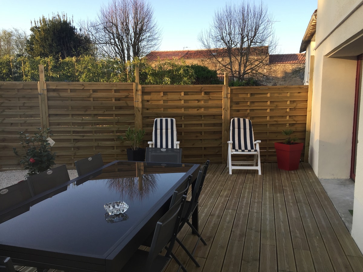 A wooden terrace with a dining table and several chairs is presented. Two lounge chairs are positioned beside a potted plant. A wooden fence and greenery surround the area, offering a sense of privacy. Natural light fills the space, enhancing the outdoor setting.