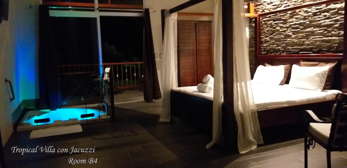 A spacious sleeping area features a four-poster bed draped in sheer white fabric, creating a serene ambiance. The jacuzzi is visible nearby, illuminated in soft blue light, while large glass doors offer access to a balcony, allowing for a connection to the outdoors.