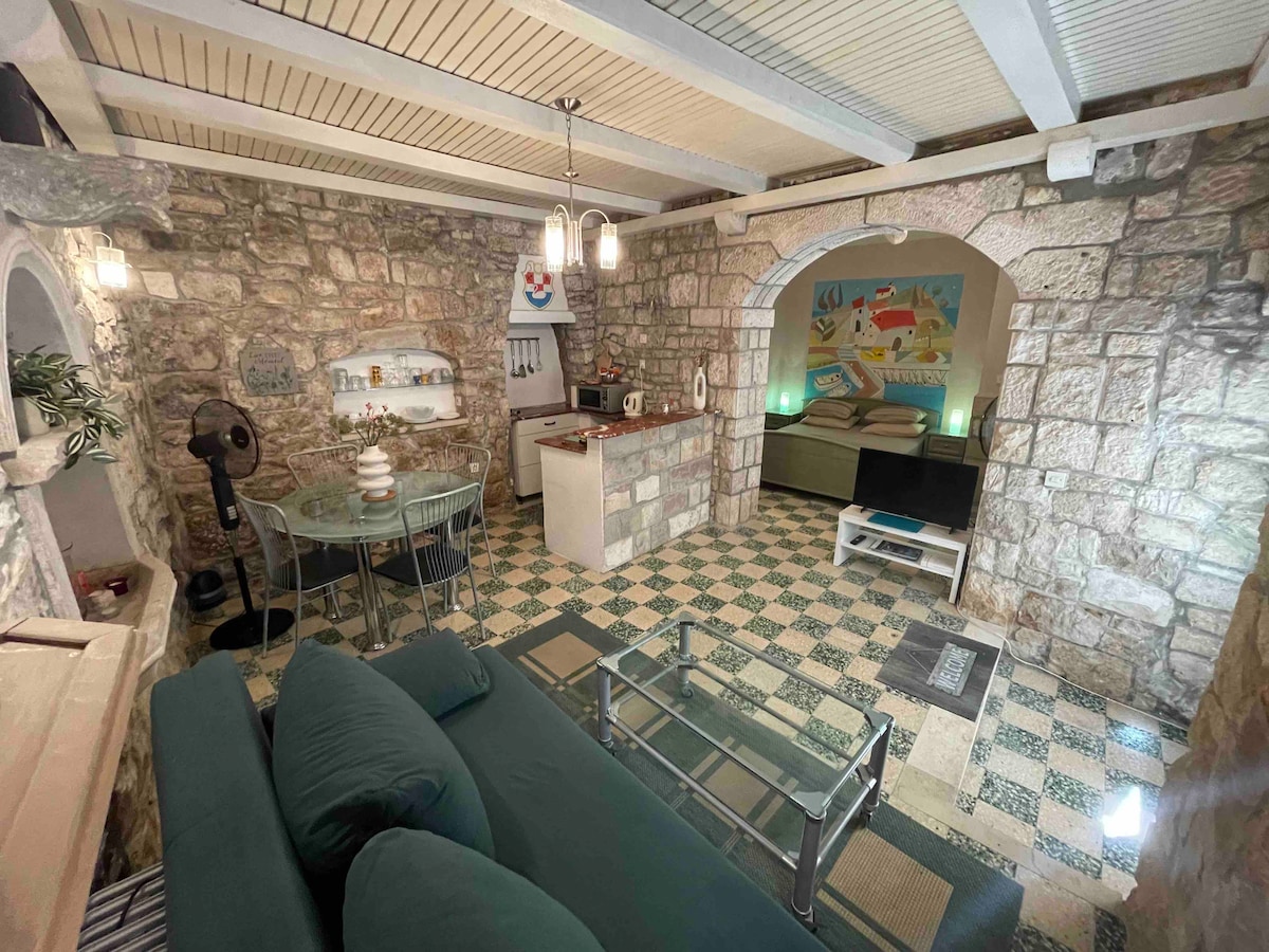 The studio features original stone walls and arches, creating a historic ambiance. A comfortable seating area is complemented by a glass table. The kitchenette, equipped with essential appliances, is situated nearby. A decorative artwork enhances the space, illuminated by ceiling lights.