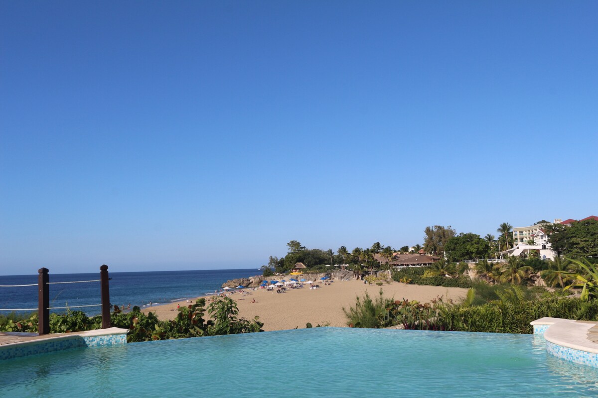 An infinity pool extends into the horizon, blending seamlessly with the sea. The beach is visible in the distance, dotted with palm trees and beach huts. A clear blue sky complements the tranquil scene, enhancing the leisurely coastal ambiance.