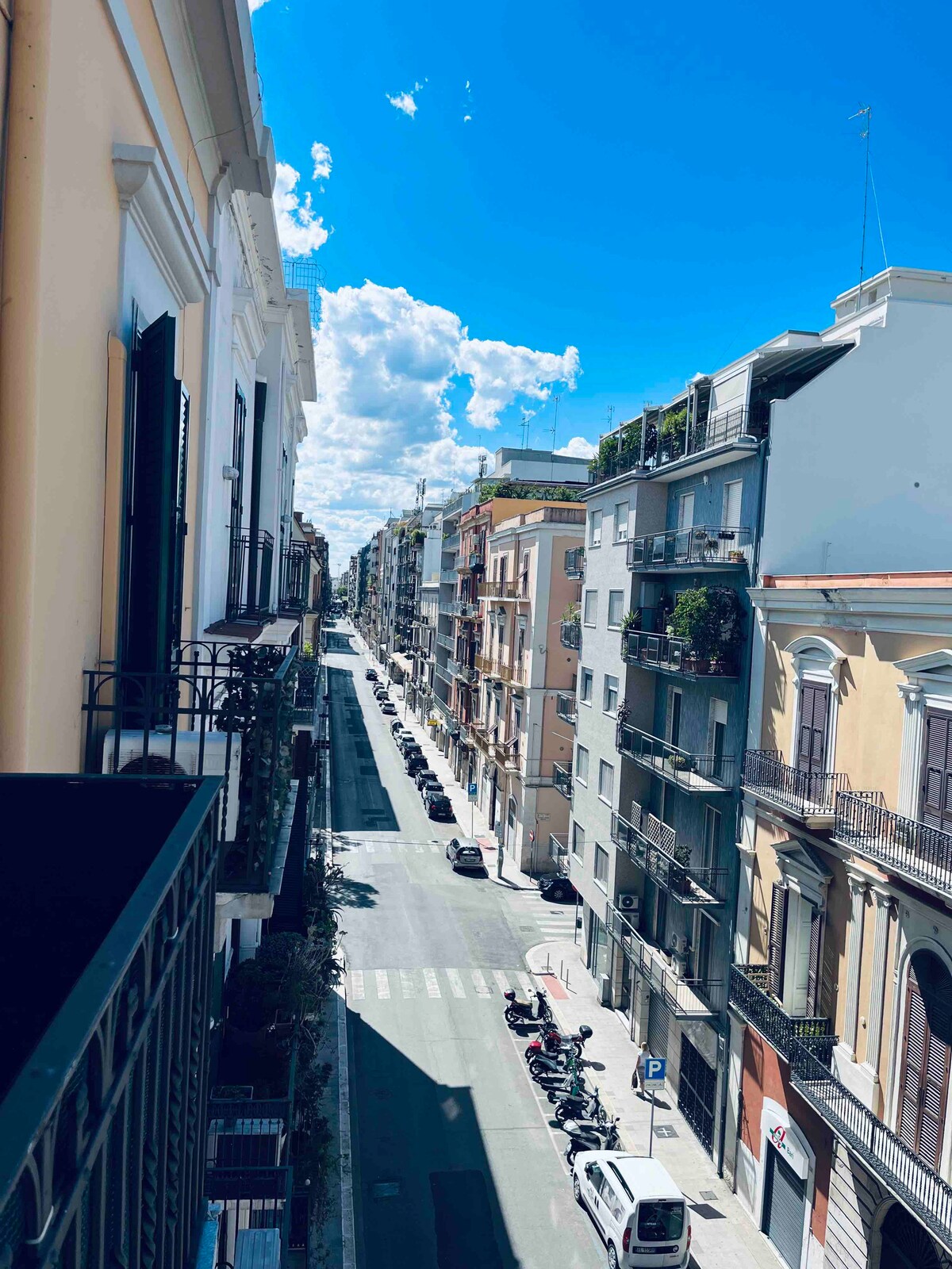 A view from the balcony captures a bright, sunny street lined with several buildings featuring balconies and greenery. The road below is marked by clear pedestrian crossings, and parked motorcycles can be seen alongside parked cars. The sky is filled with scattered clouds.