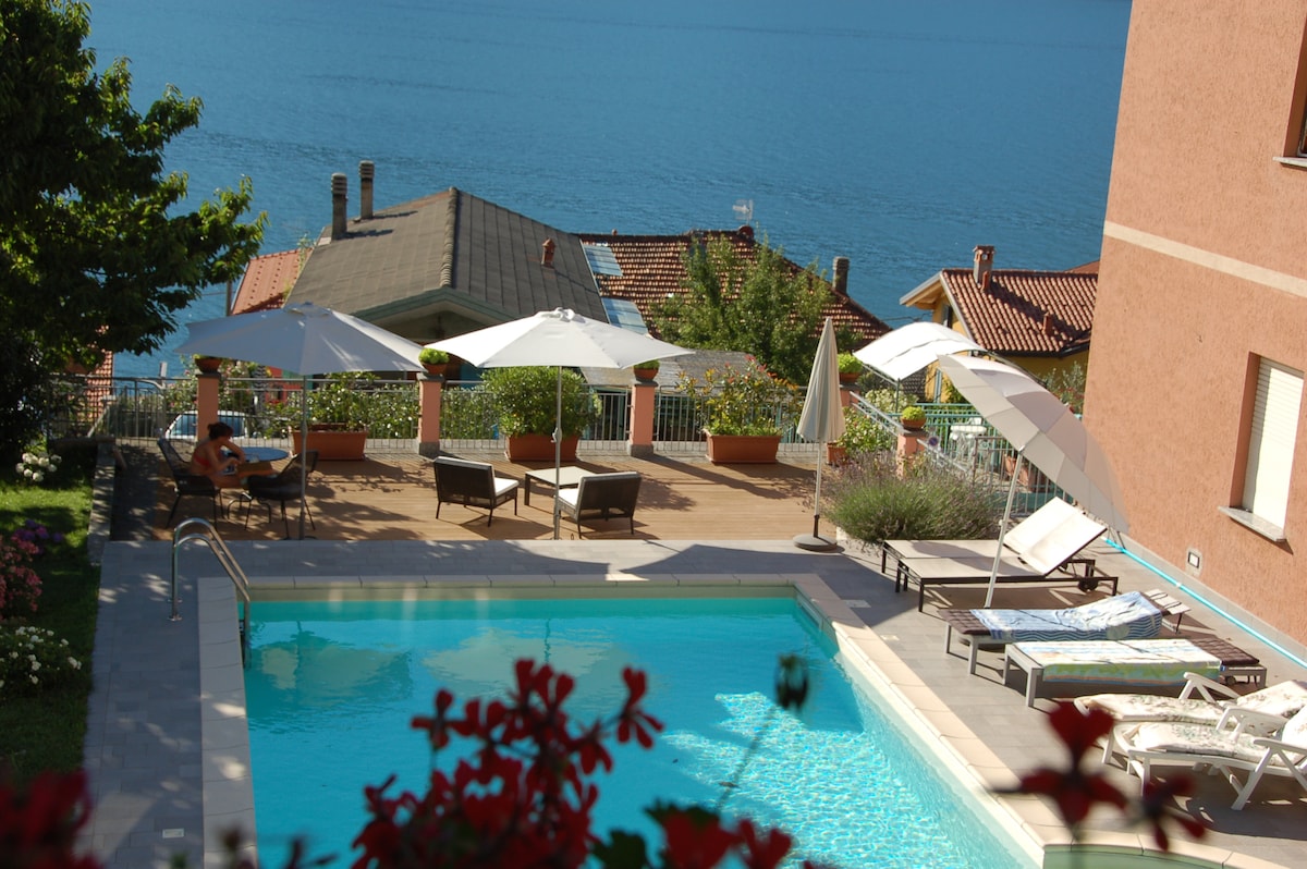 A serene swimming pool is framed by a sun terrace equipped with loungers and umbrellas, offering views of the lake in the background. Lush greenery borders the area, and the building features a warm exterior. A calming atmosphere is felt throughout this outdoor space.