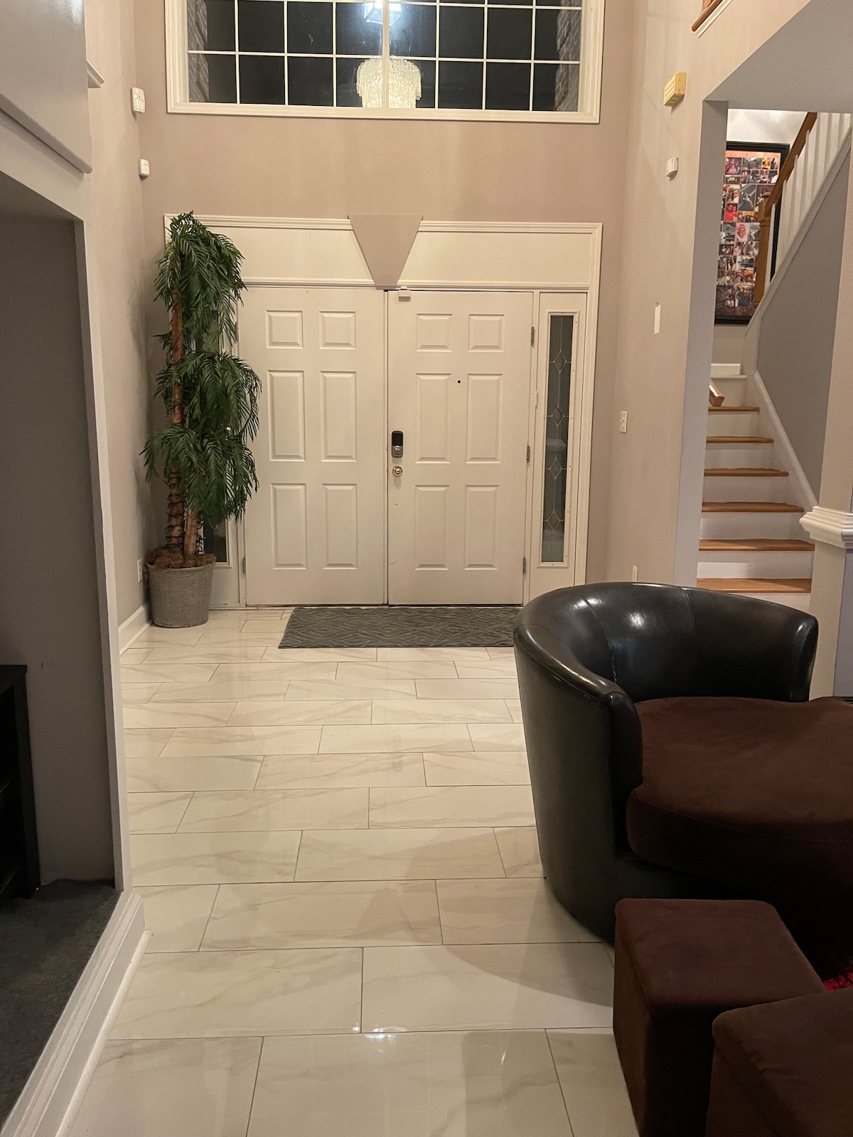 The entryway features a welcoming double door framed by a tall indoor plant in a decorative pot. Light-colored tiled flooring creates a spacious feel, while a staircase with wooden steps is visible in the background, leading to the upper level.