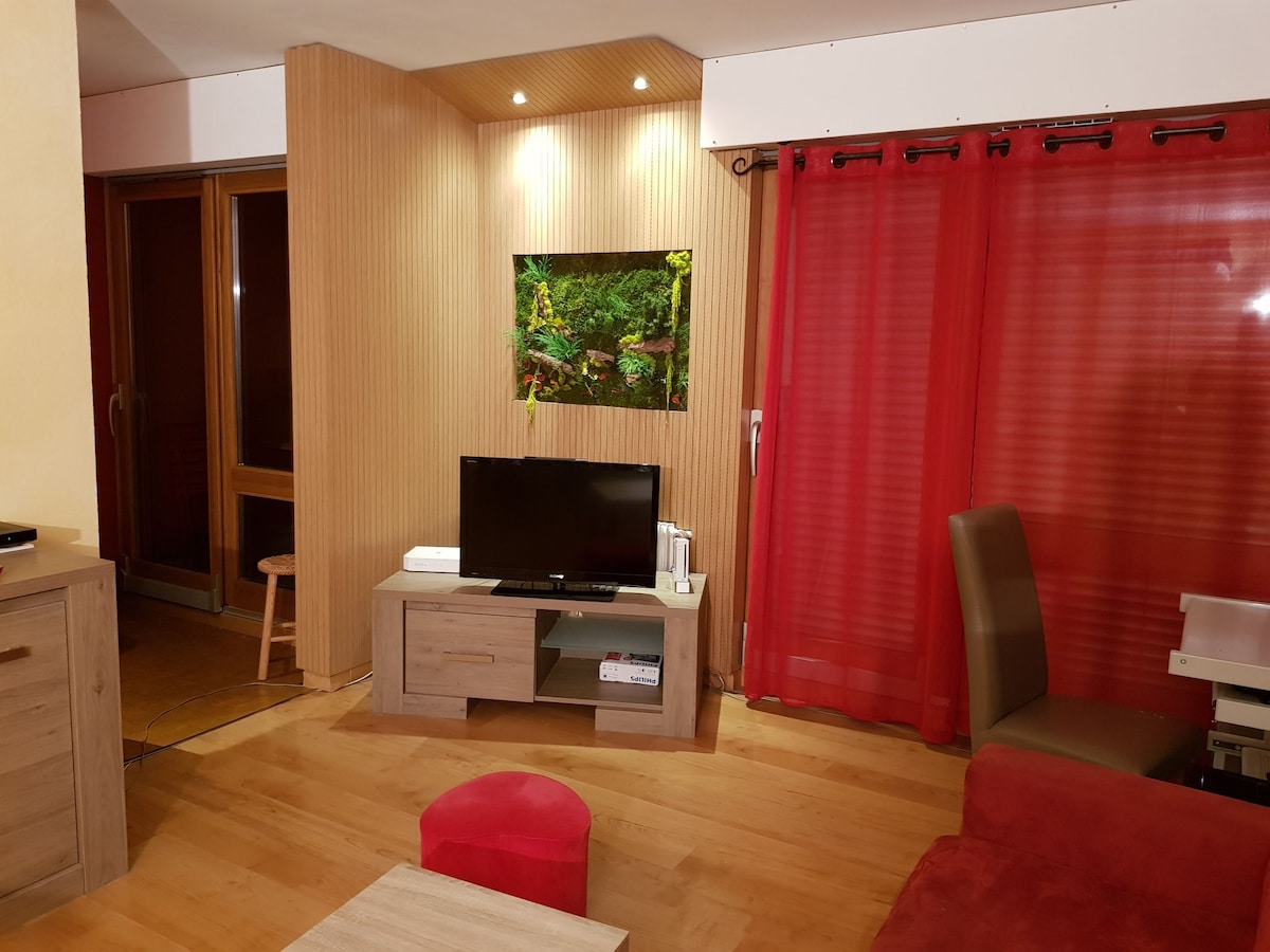 A living area is highlighted by warm wooden accents and soft red curtains. A television is positioned on a sleek media console, accompanied by a small red ottoman. Natural light enters through large glass doors, creating a relaxed and inviting space.