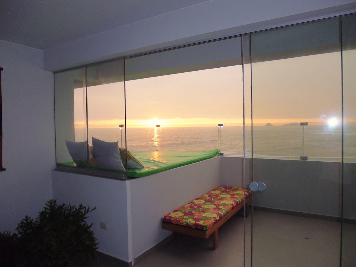 A sunlit balcony features a comfortable bench adorned with a floral cushion. The space is framed by floor-to-ceiling glass panels, offering an unobstructed view of the ocean and a stunning sunset sky. Green plants add a touch of nature to the serene atmosphere.
