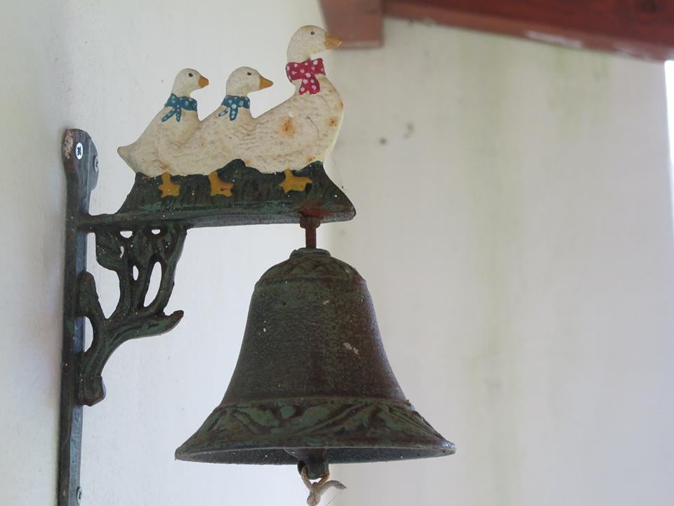 A decorative bell is mounted on a wall, featuring a whimsical design of three ducks, each adorned with colorful bows. The bell has an antique finish, complementing the muted tones of the surrounding wall.