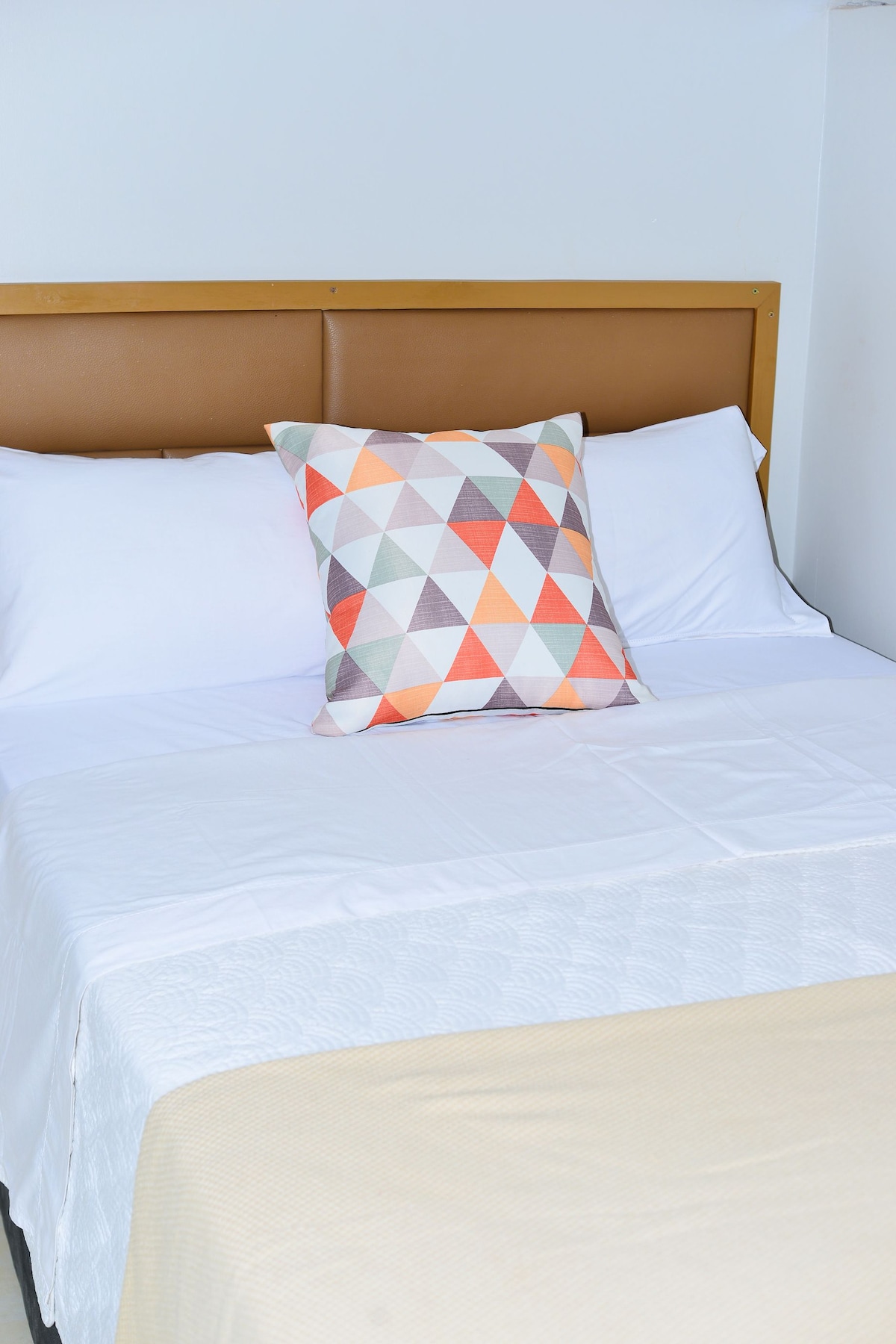 A comfortable bed is showcased with neatly arranged clean linens and a decorative pillow featuring a geometric pattern of orange, gray, and cream colors. A soft, neutral blanket is placed at the foot of the bed, contributing to the calming ambiance.