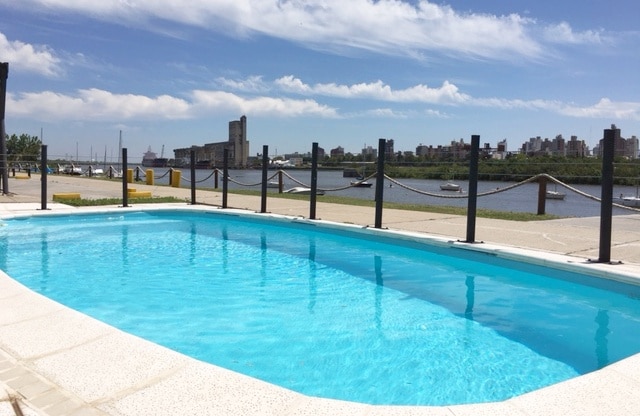 A clear swimming pool reflects the blue sky, surrounded by a light-colored deck. In the background, views of the river and city skyline are visible, including boats along the waterfront. A railing lines the pool area, enhancing safety and access.
