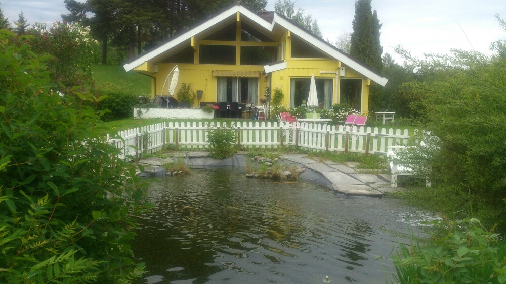 A bright yellow house with a distinct triangular roof is situated next to a serene pond, surrounded by greenery. The property features a white picket fence and outdoor seating areas with umbrellas. Large windows provide views of the peaceful landscape.