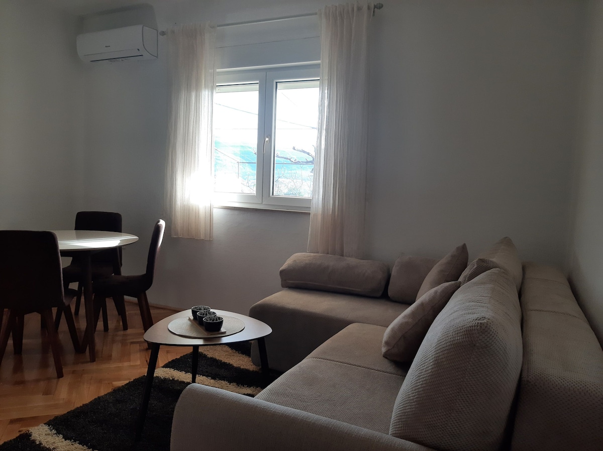 A light-filled living area features a spacious sectional sofa and a round dining table with four chairs. Soft curtains frame the window, offering views of the surrounding landscape. A small coffee table with decorative elements sits in the center of the inviting space.