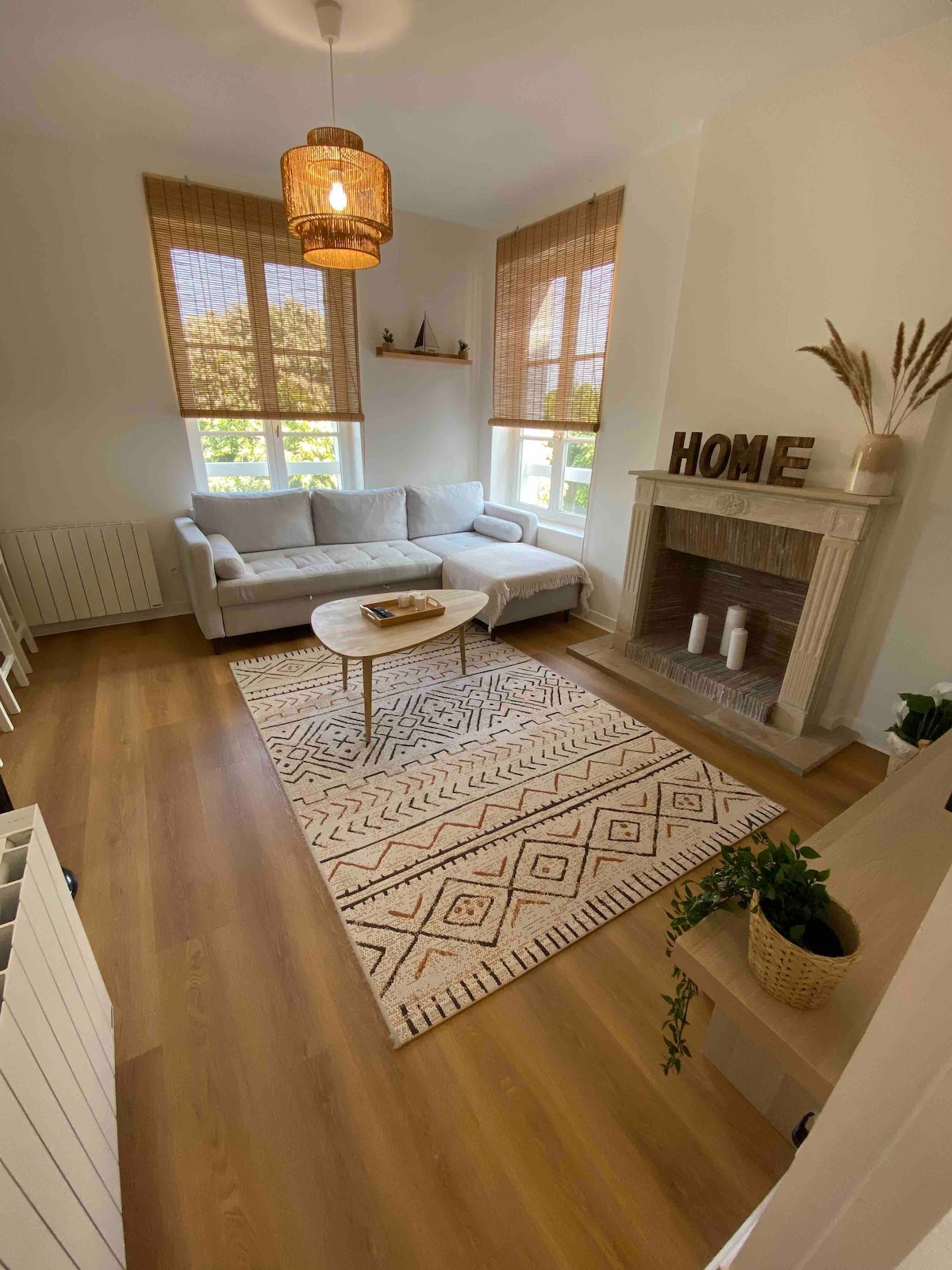 A bright living space features a comfortable sofa positioned near large windows with natural light streaming in. A cozy area rug is placed beneath a round coffee table, while decorative elements adorn the fireplace and shelves. Greenery is also present, adding a touch of nature.