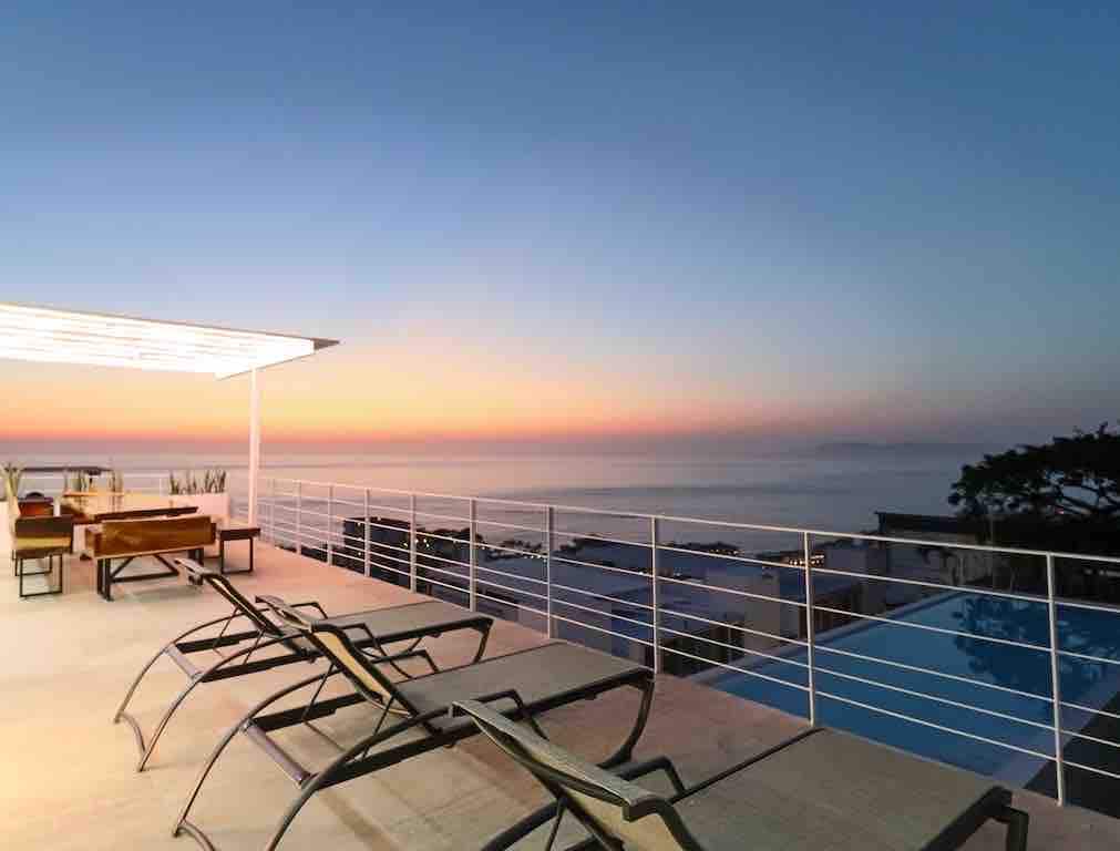 The image captures a spacious terrace featuring several lounge chairs positioned near the edge, looking out over a serene ocean view at sunset. The horizon displays a gradient of warm colors, while a covered seating area is visible in the background, creating a relaxing outdoor atmosphere.