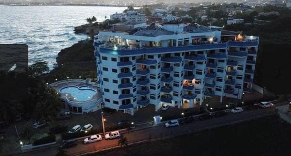 The multi-story building features a contemporary design with multiple balconies and large windows. A swimming pool is visible on the grounds, surrounded by landscaped areas and parking spaces. The proximity to the ocean is highlighted by the view of the water in the background.