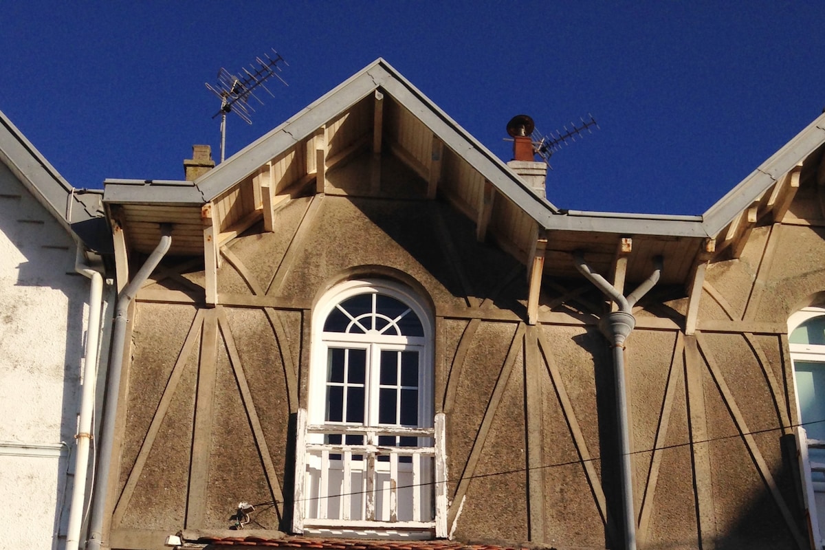 A charming architectural detail is captured, showcasing the sloped roofline of the building. A prominent gabled window with a white frame sits above a small balcony, framed by decorative wooden beams. The blue sky provides a vivid backdrop to the structure.