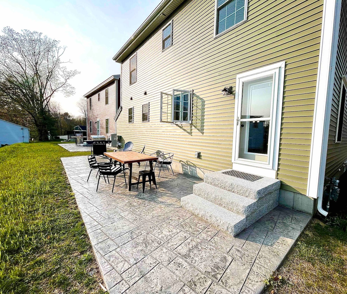 A spacious patio area features a large dining table surrounded by four chairs. Steps lead from the patio to the backyard, which is surrounded by a lawn. The exterior of the home showcases light yellow siding and several windows providing ample natural light.