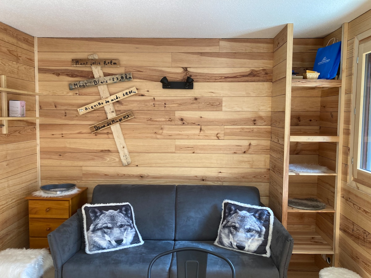 A cozy sitting area features a gray sofa adorned with two decorative pillows displaying wolf imagery. Wooden paneling lines the walls, complemented by a wooden shelf and a bedside table. Natural light enters through a nearby window, enhancing the inviting space.