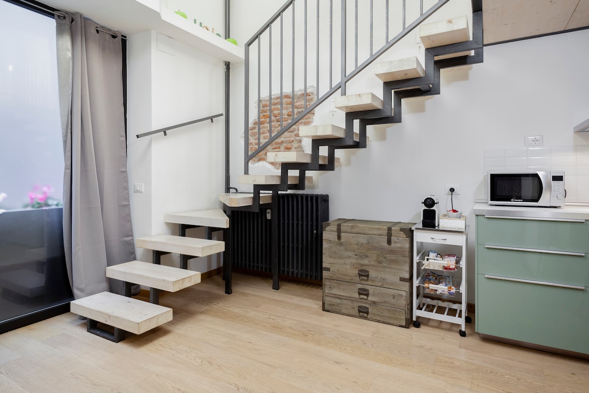 Stairs with wooden treads lead to the loft area, designed with a modern metal railing. A kitchenette is present, featuring a microwave and a green cabinet. A wooden storage chest and white utility cart are nearby, complementing the layout of the open space.