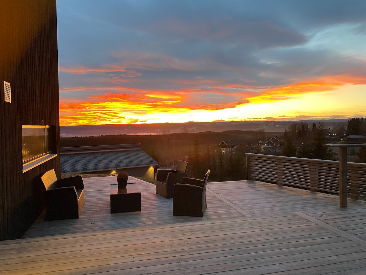 An outdoor seating area features four chairs arranged around a small table, with a striking sunset illuminating the sky in warm hues. A wooden deck stretches into the landscape, providing a view of the surrounding environment.