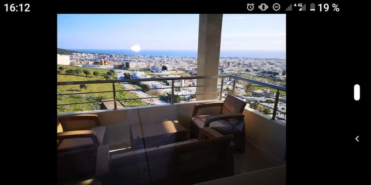 A spacious balcony is depicted, featuring a seating arrangement with four woven chairs. A panoramic view of the coastal landscape and cityscape of Bizerte is visible in the background, showcasing the blue sea and surrounding greenery.